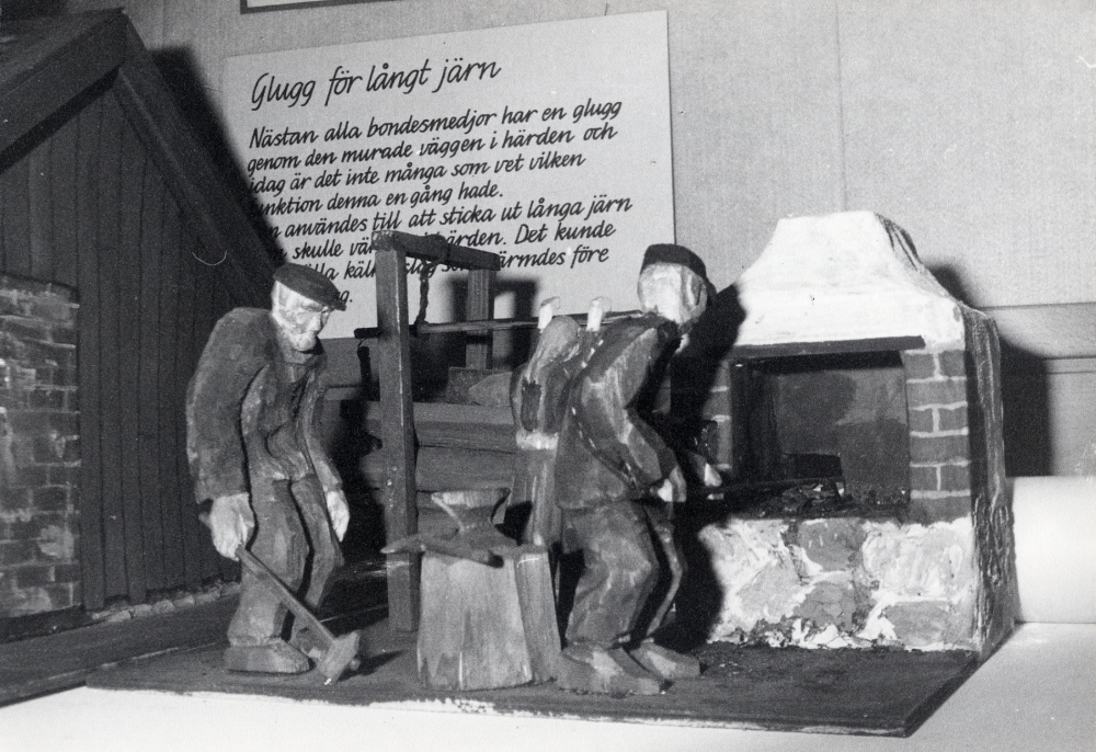 Hans Lustig har sammanställt och tillverkat träfigurer och teckningar i utställningen SMIA i Vallentunasalen. I samband med utställningen berättar han om utställningen och gamla smidestraditioner i programmet En kväll i bondesmedjan. Här syns en av träkulpturerna med text