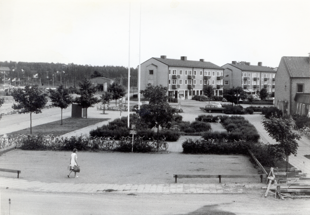 Vallentuna centrum tidigt 1970-tal, före augusti 1972. Vy över parkeringen och parken framför det gamla kommunalhuset sett från Allévägen. Södra flygeln på kommunalhuset skymtar till höger. Ett mindre vägarbete pågår på Allévägen. Längst bort ligger bostadshusen på Centralvägen med butiker i markplan.