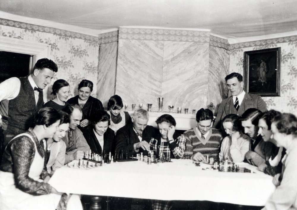 Schackklubben samlad i stora salen på Helgö gård. Runt bordet sitter fr v Alice Helgöstam, Signe Helgöstam, Sven Helgöstam, lärarinnan Elna Sandgren, Algot Laurell, Lilly Höglund, Seth Helgöstam, Sigrid Pettersson i Brogård, Helge Helgöstam, Karin Helgöstam och Helga Helgöstam. Stående fr v Holger Céwe, Edit Helgöstam, gift med Sven, Elin Johansson, barnen Helgöstams mor, Margit Helgöstam och Eskil Petterson i Brogård