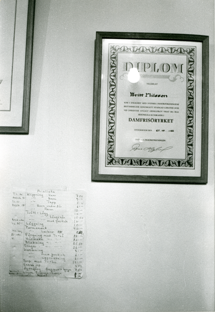 Britt Karlsson, gift med Arne Karlsson, Frösunda handel, avvecklar sin damfrisering. Britt Karlssons inramade yrkesdiplom och en prislista på väggen. Prislistan anger bland annat klippning herr 7,50 kr, dam 9 kr och klippning av barn under två år 6 kronor.
•Fotografiet ingår i bildserien ”Vardagsliv i Vallentuna kommun” av Rachael Gough-Azmier & Gunilla Blomé, Haga Studios HB.
