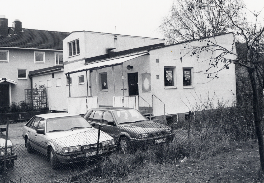 Alla fastigheterna på Allévägens norra sida mellan centrum och Mörbyvägen skall rivas. Här syns baksidan av Handelsbankens hus. Succe Färg och Tapetlager har haft sina lokaler i det vinkelställda huset men har redan flyttat till Okvista industriområde. Tre bilar står parkerade på gården