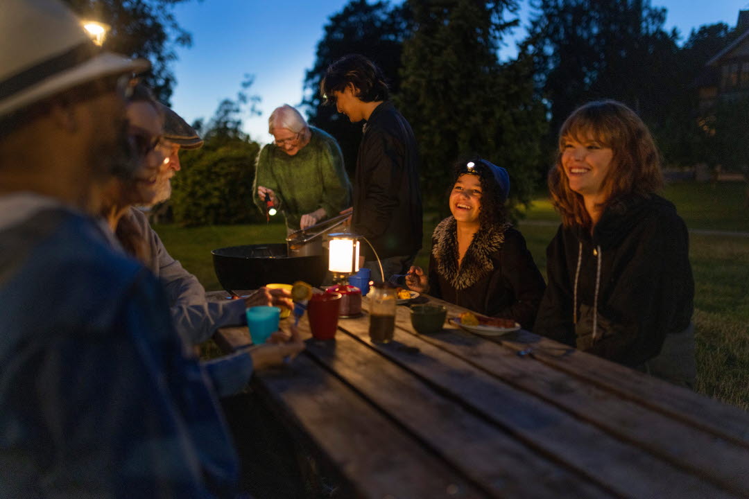 Människor i olika åldrar sitter kring ett bord utomhus i skymning. Ett par stpr i bakgrunden och grillar.