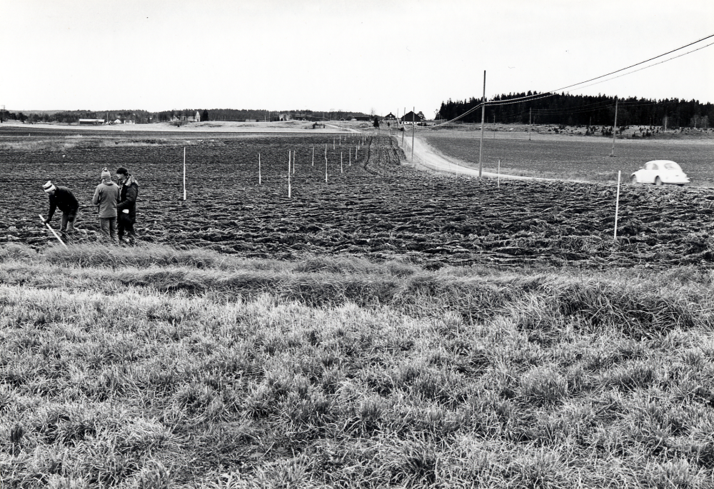 Vägen från Karby mot Vada får en ny sträckning. Markeringpinnar i åkern visar hur den nya vägen kommer att gå. I förgrunden tre män som sätter ner pinnar och i bakgrunden syns Vadaby och Vada kyrka