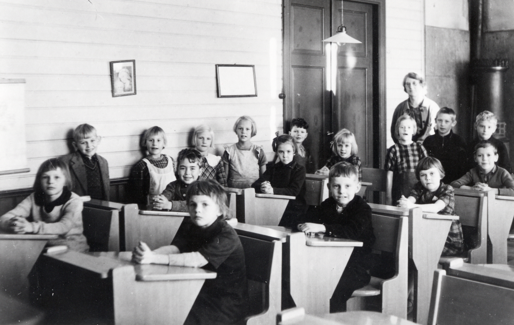 Lärarinnan Nanny Skoglund tillsammans med skolbarnen i skolsalen i Össeby-Garns kyrkskola. Pojken vid dörren heter Ingemar Lindell och ska enligt uppgift på fotots baksida ha medverkat som skolpojken Olle i filmen "Tänk, om jag gifter mig med prästen" (1941). På fotots baksida finns uppgiften 1938.