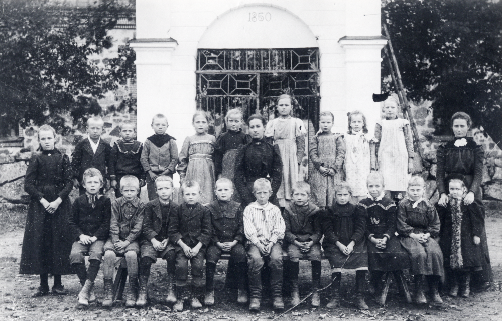 Skolfotografi utanför grinden vid Vallentuna kyrka. Lärarinnan i mitten är Anna Gauffin och t h fröken Hanna Krantz. I nedre raden nr 4 från höger är Anna Johansson  från Oppgården