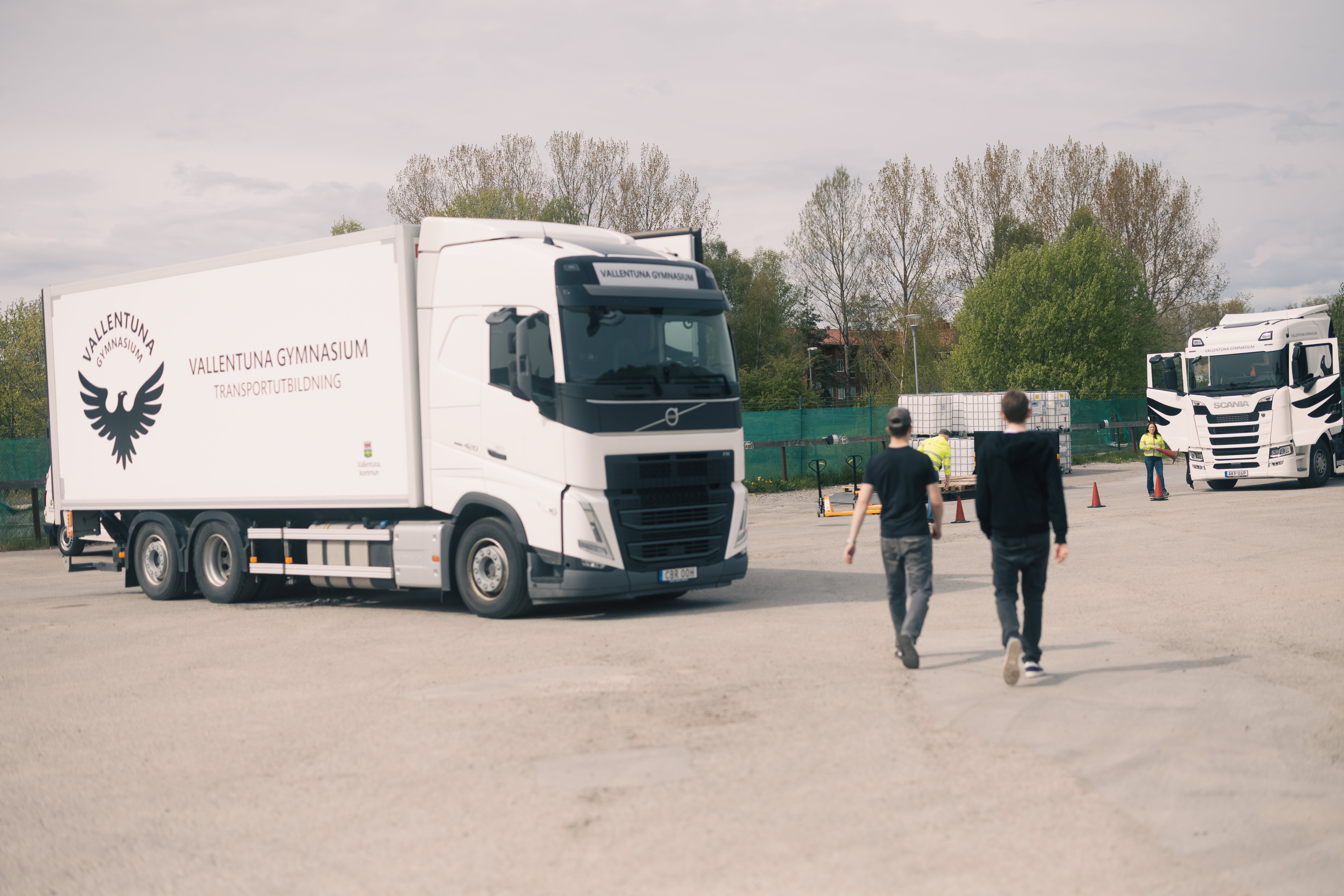 Två elever går från kameran mot transport inriktningens två lastbilar, där även läraren väntar in dem med koner utplacerade för att navigera en truck.