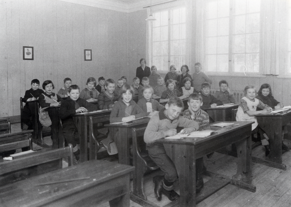 Lärarinnan Edit Lindström tillsammans med skolbarnen inne i Röda skolan i Hjälmsta.  I främsta raden längst fr t v sitter Hjalmar Linder och bredvid honom Karl-Erik Zetterlund.  Längst fram i nästa bänkrad sitter Ingrid Jansson t v och bredvid henne Eva Fredin.  Stående som 2 t h om lärarinnan är Iris efternamn okänt.