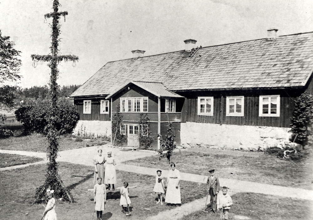 Midsommar på Helgö gård.  På gårdsplanen spelar man krocket och en midsommarstång står klädd framför huset.  Främre raden fr v okänd, Karin Johansson, Signe Johansson, Alice Johansson, Elin Johansson maka till gårdens ägare, kusinen Sven Mattsson och Seth Johansson.  Bakre raden fr v Helga Johansson och Svea Mattsson.  Barnen Johansson antog 1924 släktnamnet Helgöstam.  Svea Mattsson ser ut som en äldre kvinna, Svens mor?