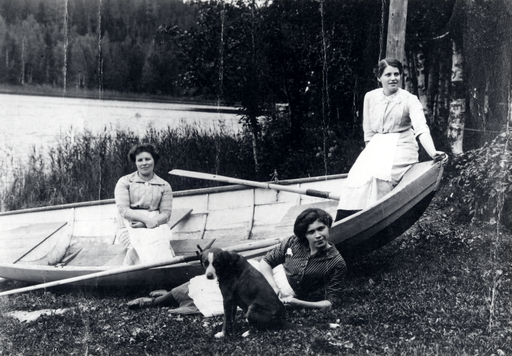 Vid stranden av sjön Tärnan i Harsbroskogen sitter tre unga kvinnor och en hund vid en båt.  Mitt i båten sitter Hulda Runnström, övriga är okända