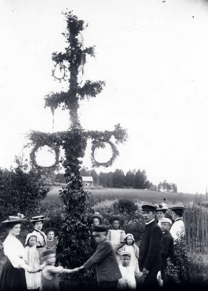 Midsommarfirande i Frösunda hos familjen Carlsson-Waldebäck. På glasplåten står antecknat: 'Vår första majstång 1908. Fr v runt majstången med början vid flickan med randiga förklädet: Alina Anjou Anna-Lisa Carlsson, Bo Carlsson, Einar Mattsson, Helge Carlsson, Zackeus Carlsson, Nils Carlsson, Lisa Barthelsson, okänd, Birgitta Carlsson, okänd, Ester Åkerlind, okänd, Louise Hermansson, besökande. Alla med efternamnet Carlsson antog släktnamnet Waldebäck.