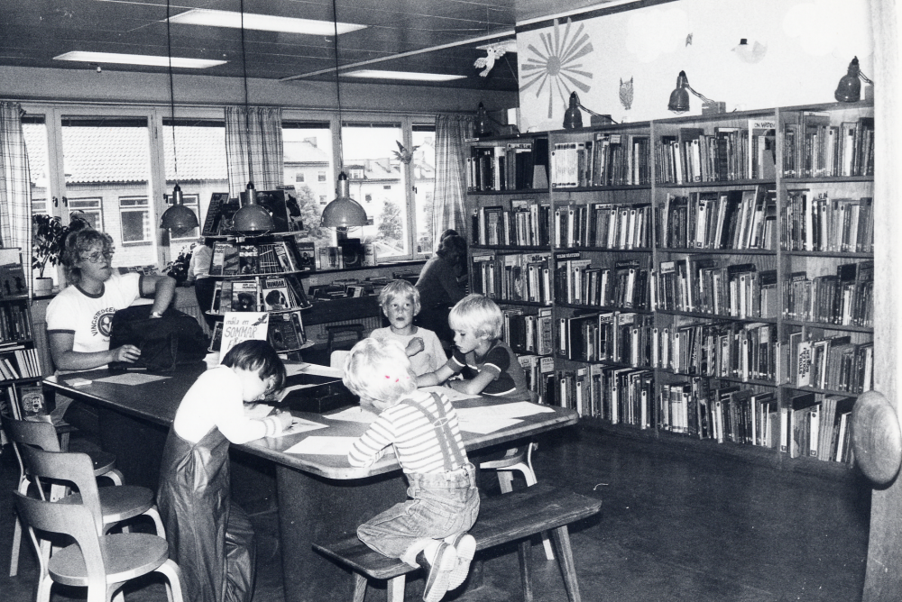Vallentuna bibliotek på Allévägen. Barnavdelningen med utsikt mot kommunalhuset.  Runt bordet sitter barn och ritar