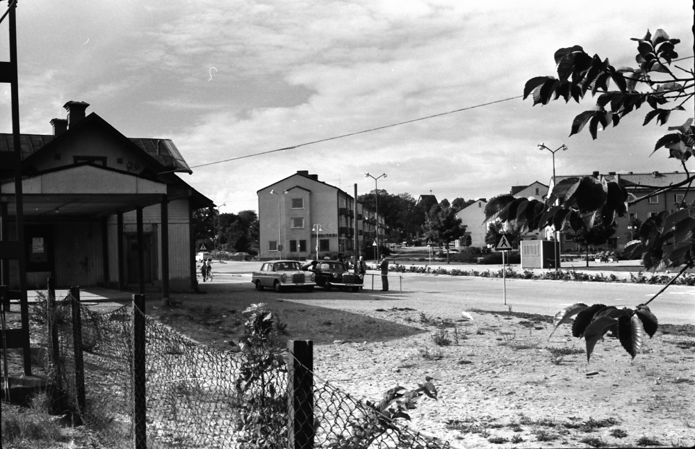 Två bilar står parkerade utanför stationshuset som syns till vänster. Mitt i bild ligger flerfamiljshusen längs Centralvägen.

•Fotografiet ingår i bildserien ”Vardagsliv i Vallentuna kommun” av Rachael Gough-Azmier & Gunilla Blomé, Haga Studios HB.