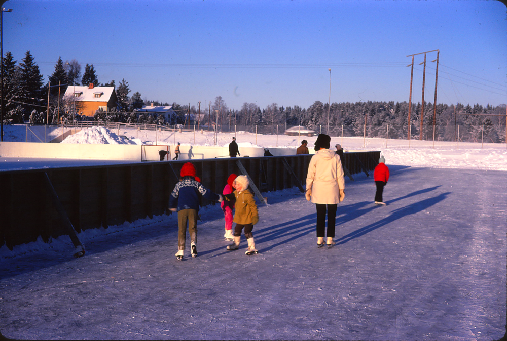 På den grusplan som i dag finns mellan Ekebyskolans idrottshall (som tillkommit i sen tid) och Norrgården fanns en rink där det spolade is vintertid. Flickan i den bruna jackan och vita mössan är Åsa Nilsson (gift Breimo)och kvinnan i den vita jackan och bruna mössan är Margareta Nilsson. Teknikvägen går i mitten av bilden, den gula villan finns än i dag och Hammarbacksskolan syns i bakgrunden. VInterbild. Februari.