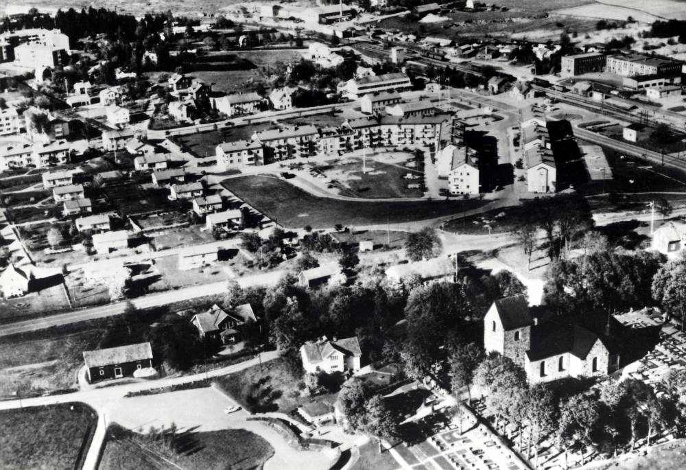 Flygfoto  över det bebyggda  Prästgårdsgärdet  med  Vallentuna kyrka  i förgrunden. I bildens mitt bostads- och affärshus på  Centralvägen  med  Vallentuna järnvägsstation  t h  Maskin-Tellus  t v enfamiljshus. Högst upp i bildens mitt  Vallentuna tegelbruk