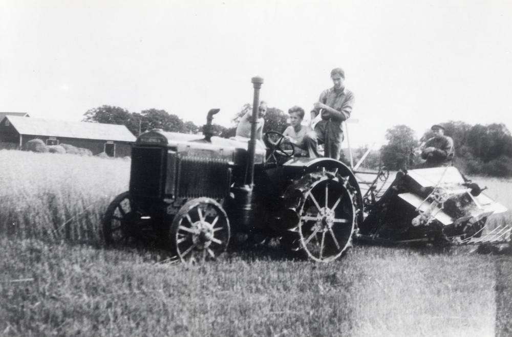 Skördebindare vid Frösunda prästgård. På traktorn sitter prästsönerna Karl-Erik, Lars-Olof och Jan-Hans Sjöberg. Prästgårdsarrendatorn G E Carlsson sitter på bindaren. I bakgrunden syns ekonomibyggnader vid Prästgården