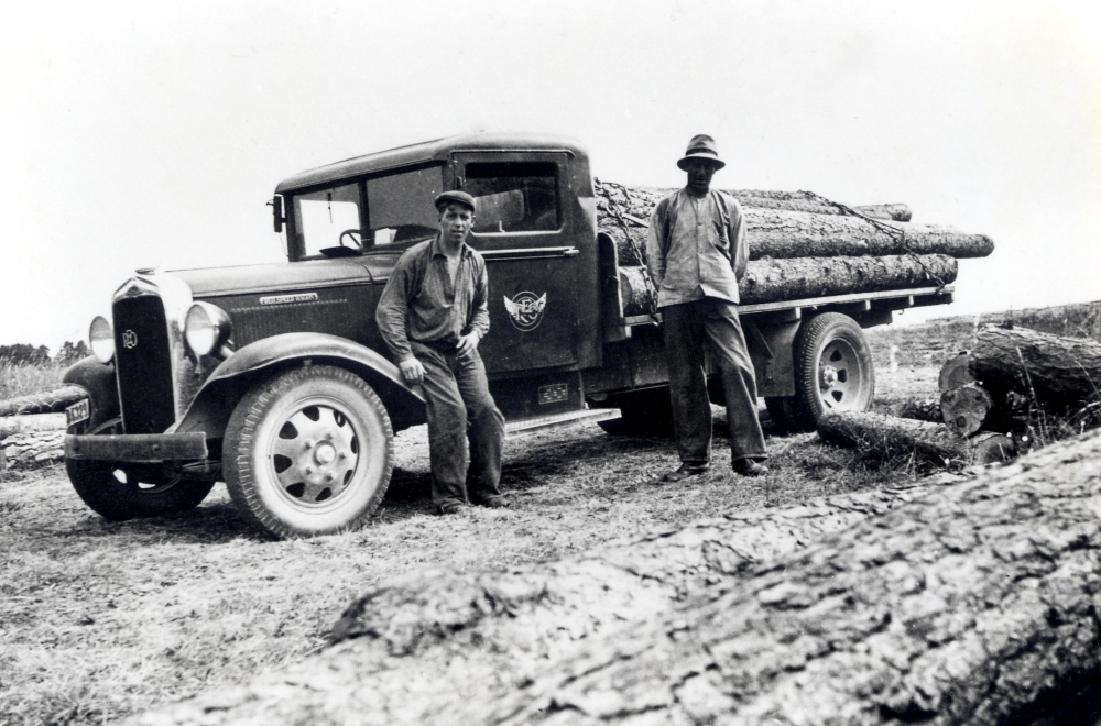 Timmerbil, ev vid sågen i Husa.  Bilen tillhörde ev Alstermarks åkeri. T v står Eric Ericsson och t h ev Fritz Johansson.  Eric Ericsson startade eget åkeri ca 1937.  Efter hans död drev sonen Sven Eric och mågen Tord Lundgren företaget vidare fr o m 1 januari 1973.  Åkeriet upphörde 31 december 1995.