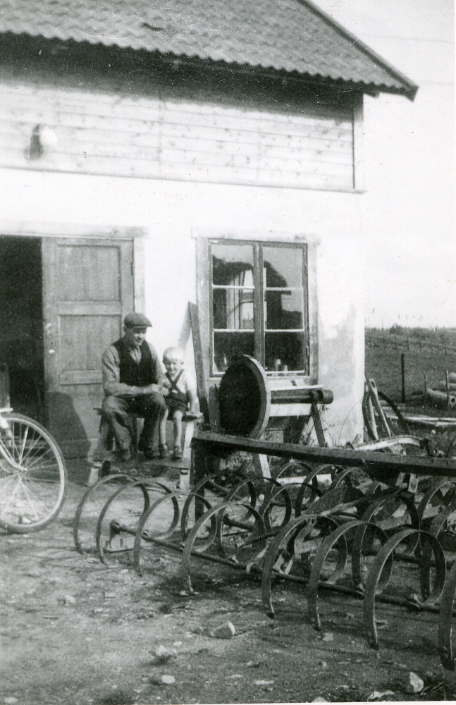 Seth Axén vid smedjan mitt emot macken i Angarn.  Bredvid Seth sitter sonen Åke, född 1946 eller möjligen sonen Bengt född 1942.  I förgrunden står en harv