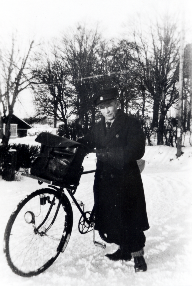 Helge Karlsson, "Post-Kalle", delar ut posten i Angarn.  Under de kalla krigsvintrarna på 1940-talet, var det ett hårt arbete att cykla runt och dela ut post på landsbygden.  I bakgrunden t h skymtar Angarns kyrka.