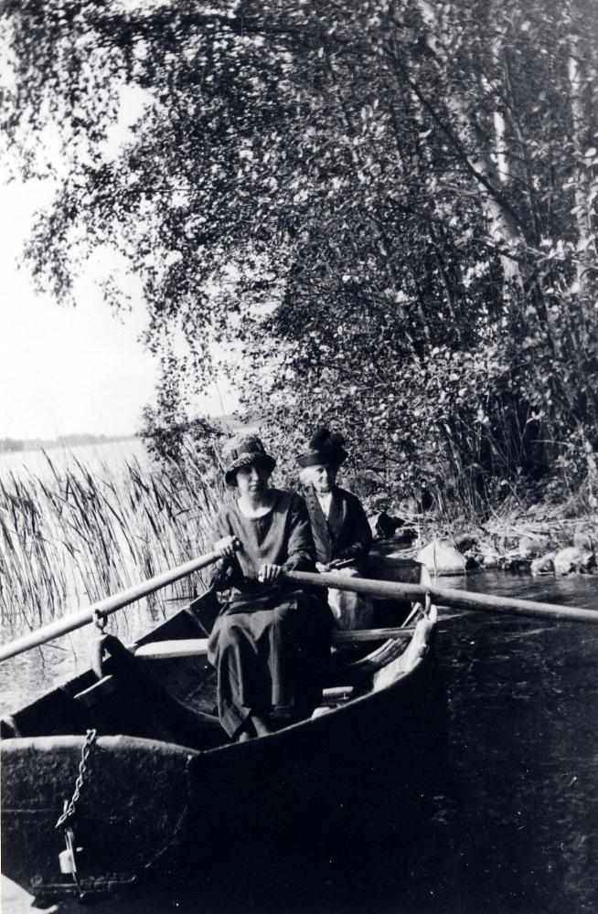 Märta Johansson  från  Oppgården  sitter vid årorna i en  roddbåt  på  Vallentunasjön  På aktertoften sitter hennes mamma  Tekla Johansson  iklädd  hatt med plym  Här har man gjort strandhugg vid  Bällsta strand