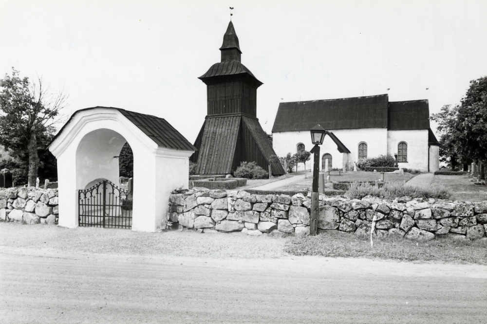 Orkesta kyrka i förgrunden grindar med portal och stenmur. T v Klockstapeln