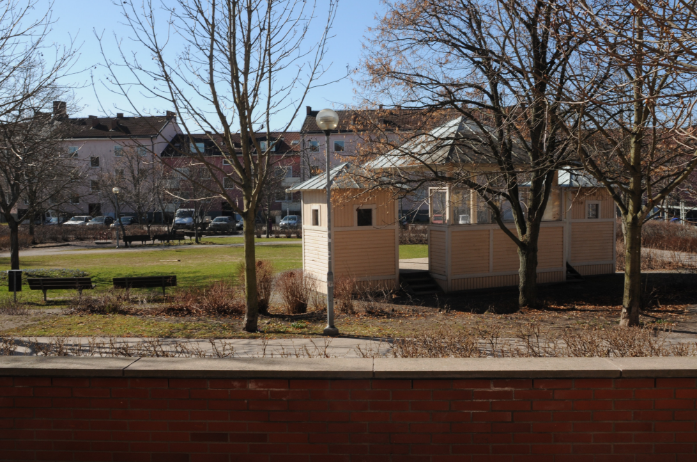 Kommunalhusparken i Vallentuna centrum med scenen till höger, sett från Allévägen. I bakgrunden syns flerfamiljshusen på Skördevägen.
