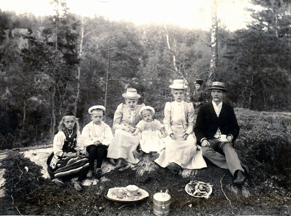Någon gång i början av 1900-talet är en familj på utflykt i naturen. Området verkar ha en del berg i dagen. På marken står en kaffetermos, ett fat med kakor och en bricka med socker och mjölk.