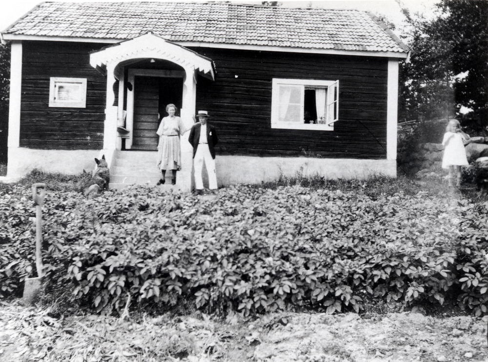 Nygård, torp under Rävsta gård (med fastighetsbeteckningen 1:5).  Framför huset  vid farstubron står åkare David Wesström och hans hustru, t h står dottern.  Familjen hyrde Nygård som sommarbostad.  Vid trappan sitter också en hund.  Framför trappan finns ett potatisland och en spade