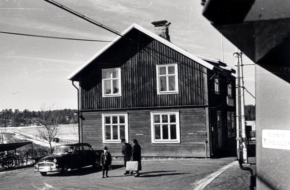 Ekskogens järnvägsstation.  Med sin Vedette Versailles, årsmodell 1956 med registreringsnummer B 52920,  hämtar Manfred Ekholm från Ekskogens gård hyresgästerna i gårdens södra flygel.  Manfred står i mitten, pojken heter Bengt-Åke Hallsten och damen Aina Hallsten  Fotograferat från tåg 16. Ekskogens stationshus revs sommaren 1963 av Nils-Erik Rignér och dennes far. Huset var i bra skick och virket kunde återanvändas. Stationeshuset hade varit utan tågklarerare sedan 1962?..