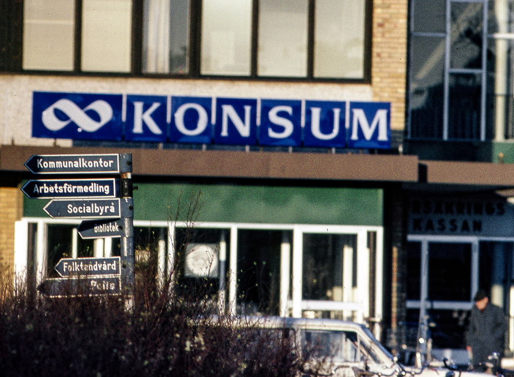 Konsums fasadskylt på Torghuset, tidigare kallat "Konsumhuset". I förgrunden står en vägvisare med skyltar; Kommunalkontor, Arbetsförmedling, Socialbyrå, Bibliotek, Folktandvård, Polis.
Bildserie: Vallentuna på 1960-talet.