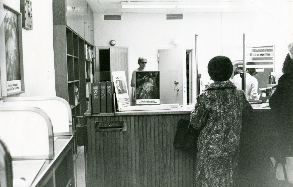 Interiör från postkontoret på Centralvägen. En kund står framför glasskärmen vid disken, personal syns där bakom.

•Fotografiet ingår i bildserien ”Vardagsliv i Vallentuna kommun” av Rachael Gough-Azmier & Gunilla Blomé, Haga Studios HB.