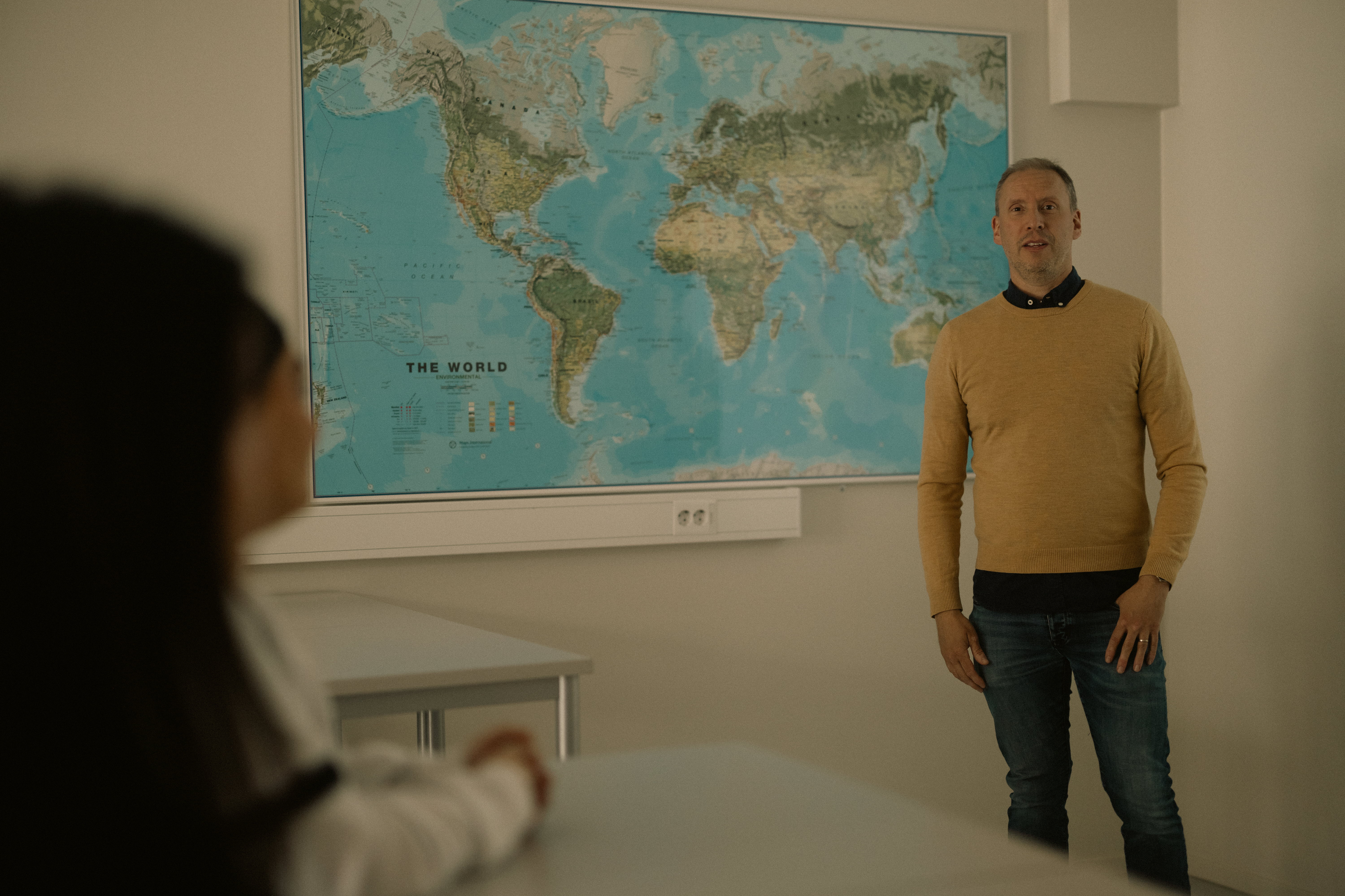 Lärare står framför en världskarta och håller lektion framför eleverna som sitter vid sina bord och lyssnar.