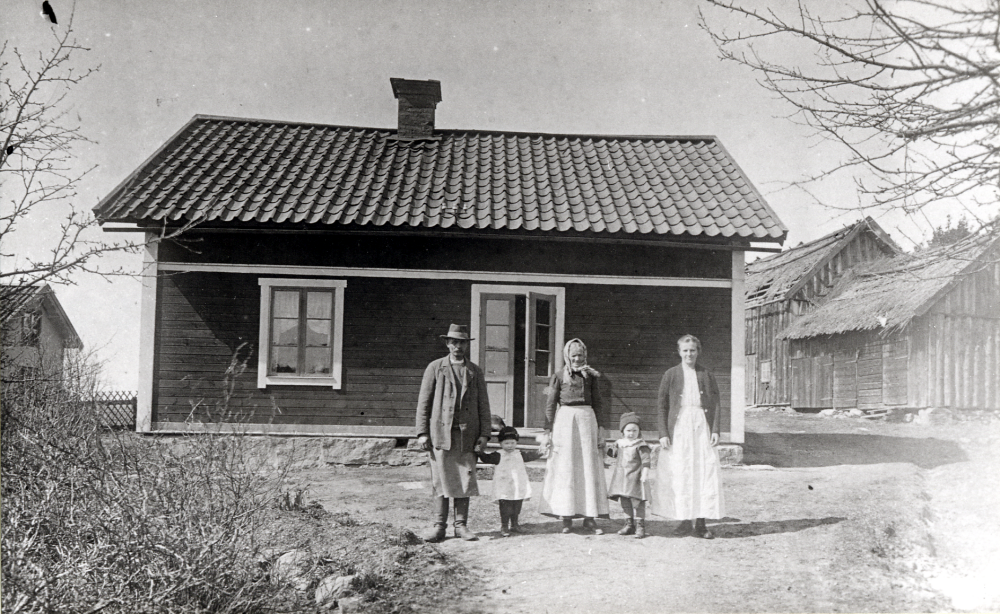 Familjen Plomgren utanför huset i Ubby.  Fr v  "gubben" Plomgren med hustrun Tilda och sonhustrun Frida, gift med det äldre parets son Valle.  Barnen heter Maja och Georg.  En syster till Tilda, som hette Emma, bodde på vinden.  Plomgren hade en häst.  Frida kom från Klippinge i Närtuna.  Valle var "en slarver" och den  unga familjen hade svårt att leva på vad gården gav.  Valle hade en syster Vera, som byggde nytt hus i Ubby på denna byggnads plats.  Ekonomibyggnaderna t h om boningshuset  har  trasiga halmtak. T v syns sannolikt Ubby 1:21 Solhem, 1952 ägdes det av fd fabrikören Wilhelm Hellgren.