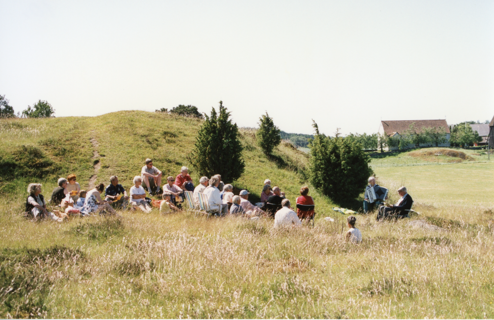 En grupp besökare har tagit plats i gräset vid Vada sjökullar under en friluftsgudstjänst.
Längst till höger i bild sitter Nils-Göran Enggren, kyrkoherde em och IA Arhusiander som spelar dragspel. I bakgrunden syns Vada kyrka.