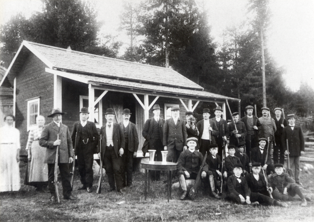 Medlemmar i Vallentuna skytteförening utanför  gamla paviljongen vid Fågelsången. Mannen i bildens mitt med benlindor är Carl Bornholm, med slokhatt längst t v står Henning Henningsson.  Iklädd kubb bredvid pojken med hunden står Ferdinand Lindström.  På bordet står två pokaler.