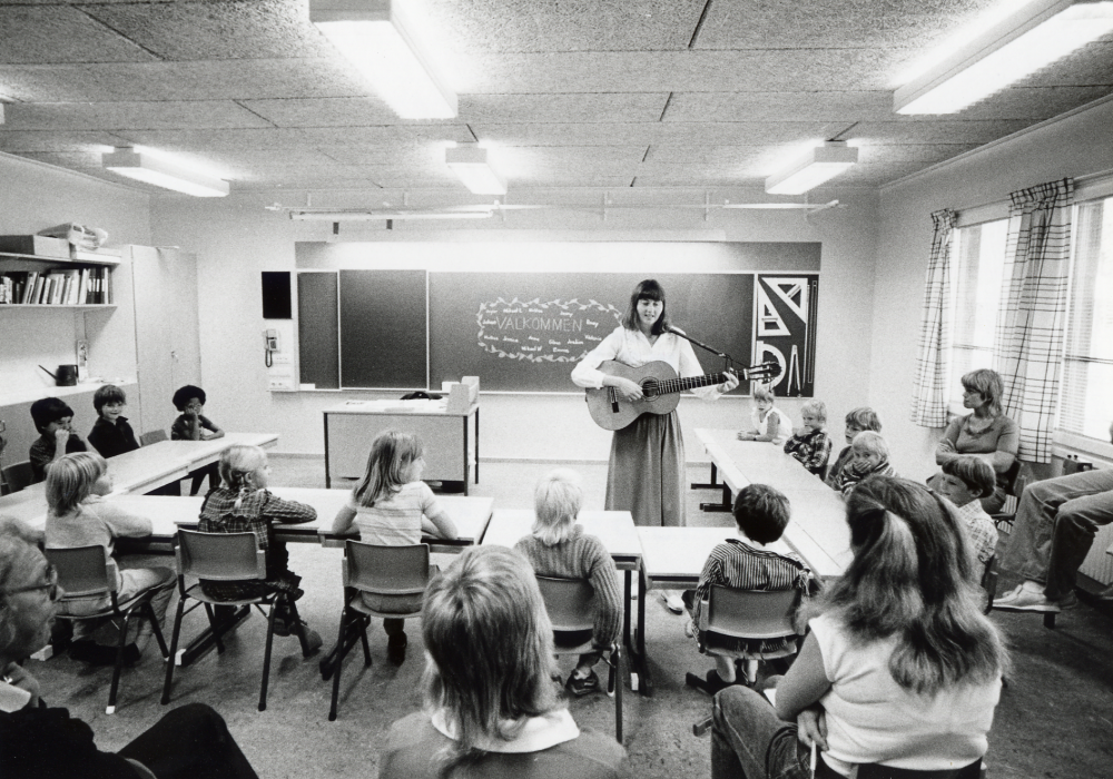Skolbarnen  sitter i klassrummet första skoldagen i nya Kårstaskolan höstterminen 1982.  Lärarinnan ?????? spelar gitarr.  Några föräldrar har också fått följa med in i klassrummet