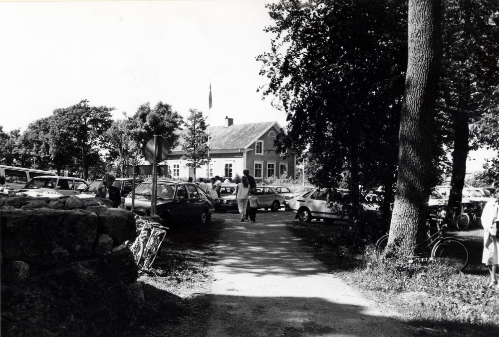 Många människor är på väg till midsommarfesten på Kyrkängen nedanför Vallentuna kyrka. Parkeringsplatsen vid kyrkan är full av bilar. I bildens mitt syns Klockargården