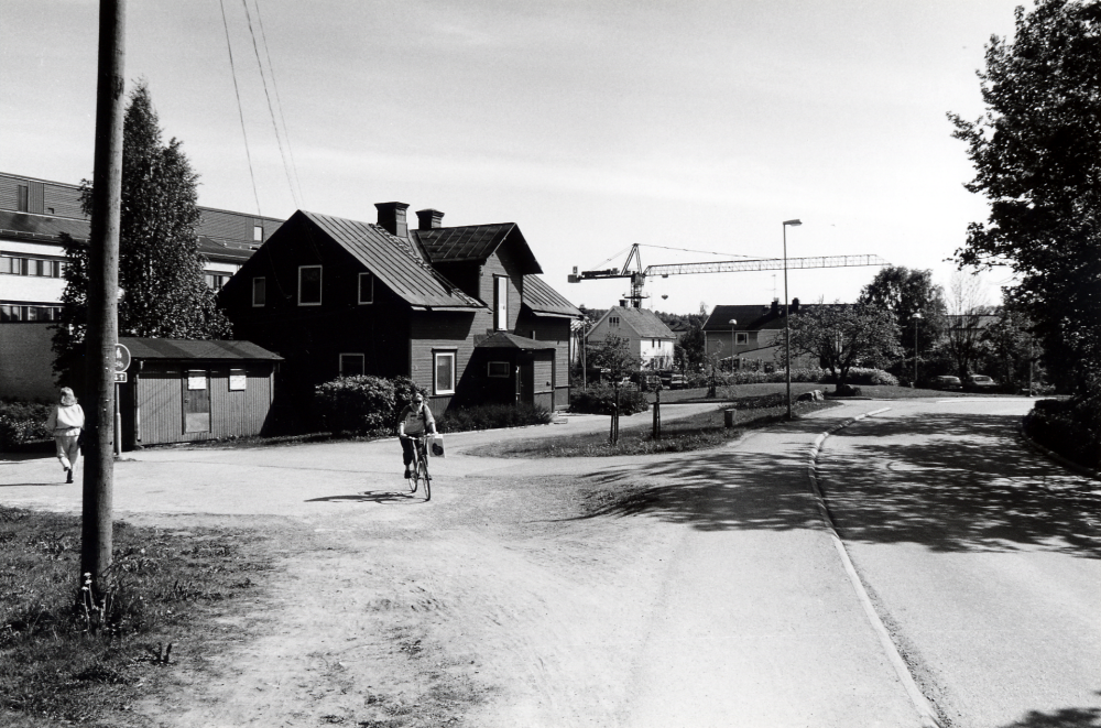 Vallentuna centrum sett från Mörbyvägen med gångvägen till centrum t v, syskonvillan, och Mörbyvägen ner mot Allévägen. Centrumhuset Tärningen skymtar bakom syskonvillan och bebyggelse längs Allévägen nedanför byggkranen. Den är från det nya kommualhusbygget