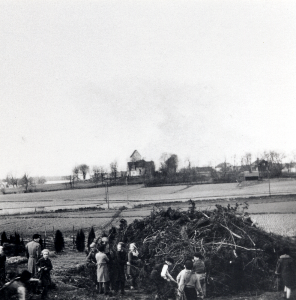 Skolbarn samlar ris och skräp till årets valborgsmässoeld nedanför Hjälmsta skola.  I bakgrunden syns fr  Sjövillan, kyrkan, Klockargården, kyrkoherdebostaden och arrendatorsbostaden