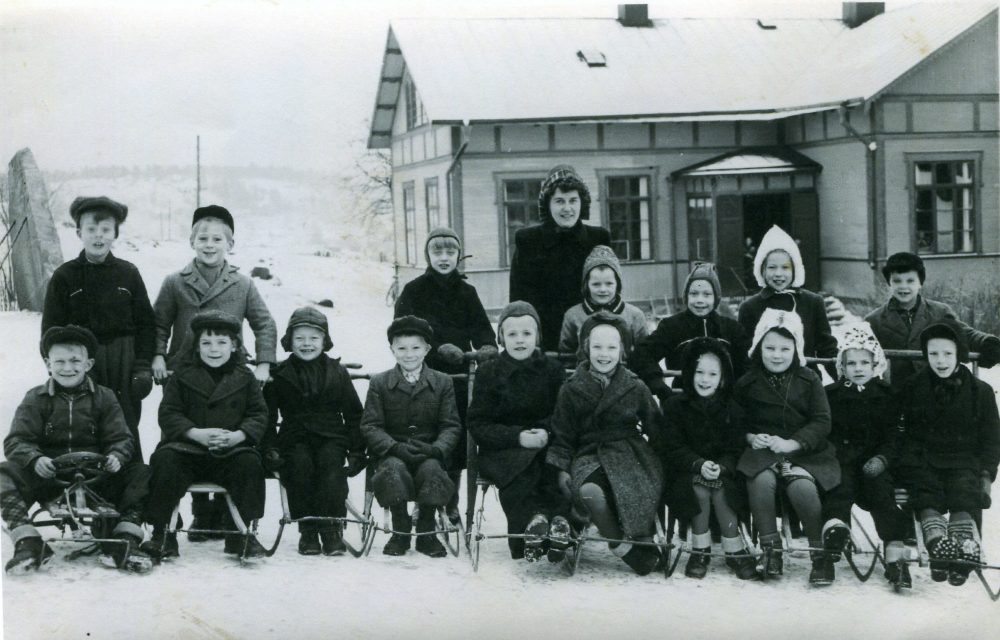 Lärarinnan Gunborg Sjöström fotograferas utanför skolan tillsammans med barnen i Össeby-Garns folkskola. Birgitta Lindell klass 2 sitter på sparken trea från höger.
Namn textat på baksidan, från vänster bakre raden, Yngve A, Lars R, Arne J, fröken Sjöström, Gunilla E, Britt A, Ragna A och S Hägg?.
Främre raden, Olle P, Göran ?, Lars G, Leif J, Ing? K, Eivor J, Margareta B, Birgitta Lindell, Britt N och Hans P. Barnen är vinterklädda och raden längst fram sitter på sparkstöttingar och kälkar. Snö täcker skolgården och skolhusets tak. Årtal saknas.