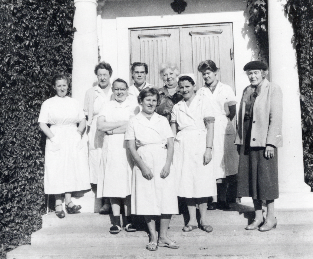 Elsa Tiberg, kallad "Fröken",  klädd i mörk hatt med del av sin personal på trappan vid Bällsta gård - Vallbostrands vårdhem. Elsa Tiberg hade först pensionat på gården, 1928-1933 och sedan vårdhem för vuxna med utvecklingsstörning.  Fr  v står Carla Lilja som var husmor de sista 10 åren, Fru Johansson, okänd, Svea Gullmes, Margareta Bertilsson, Esster Granberg tvätterska, okänd kokerska, samt Elsa Tiberg okänd   Vårdhemmet hade ca 13 anställda.
Jfr 90 04 00: 004