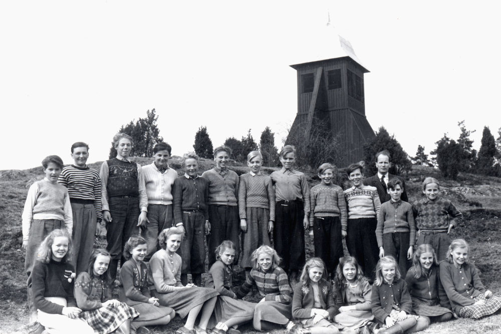 Storskolan i Kårstaby, klass 5-7.  Läraren Halvar Hedler med skolbarnen vid klockstapeln.  I övre raden fr v står: Arne Johansson Henriksborg, Björn Helgöstam Tjusta, Sven Kraft Ledinge, Bernt Sundberg Henriksborg, Roland Andersson Grönlund, Per-Olof Karlsson Solbo, Sven-Olof Öhman Karlsro, Holger Ljungstedt Kvarnhagen, Gunnar Jansson Nibble, Harry Bergkvist Lövstalund, magister Halvar Hedler, Lennart Elgebrandt Sunnarby och Bjarne Ritzén Kårstaby.  Främre raden fr v: Ingegärd Enderberg Tallsäter, Irene Lundin/Jansson Bertilsberg, Stina Andersson Ekskogens station, Marianne Andersson Kårstaby, Kerstin Lillje Sunnarby, Siv Jansson Backa, Siri Jansson Backa, Ingegärd Söderström Skuggan, Margit Karlsson Backa, Eivor Kraft Ledinge och Majvi Karlsson Sofiero .  Ej med på kortet: Karl-Erik Berglund Bersjödalen, Anders Ohlsén Malmberget, Bo Sjöholm Kyrkskolan, Bo Allan Berglund Berghem, Karl-Erik Karlsson Nystugan, Karl-Axel Jansson Salby och Gun Jansson Ledinge