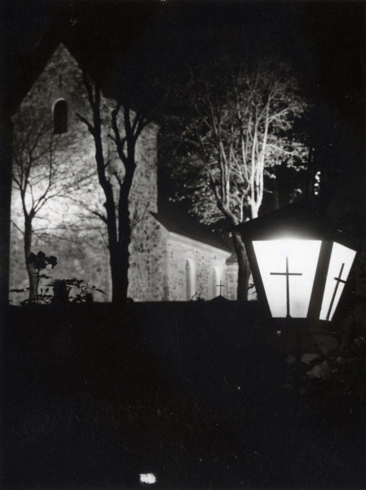 Vallentuna kyrka i strålkastarsken sedd från sydväst mot tornet med en gravlykta i förgrunden