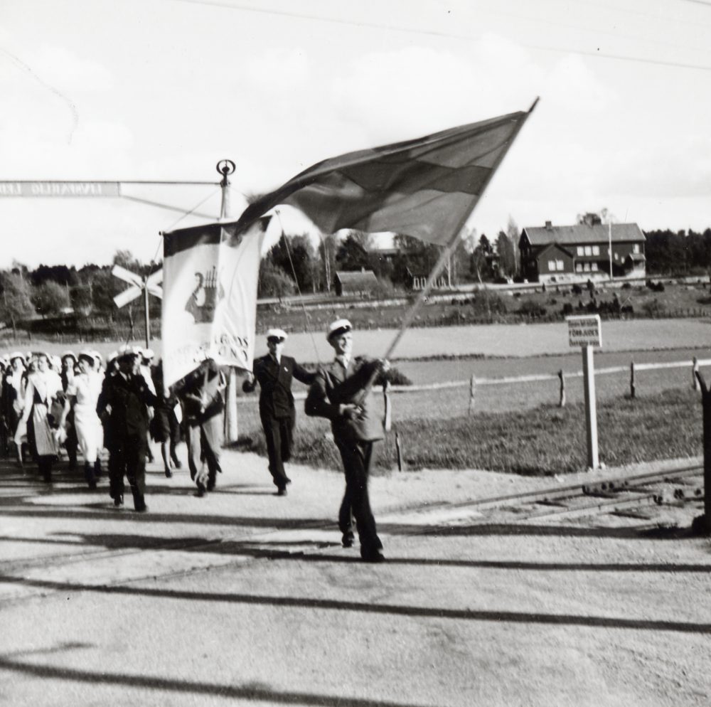 Östra Upplands körförbund  på väg mot  Vallentuna kyrka  för att hålla konsert i kyrkan.  Fanbärare  är körens ordförande  Arne Lindström  Kören vandrar över landsvägens korsning med  Roslagsbanan. I bakgrunden t h syns  Hjälmstaskolan   Röda skolan