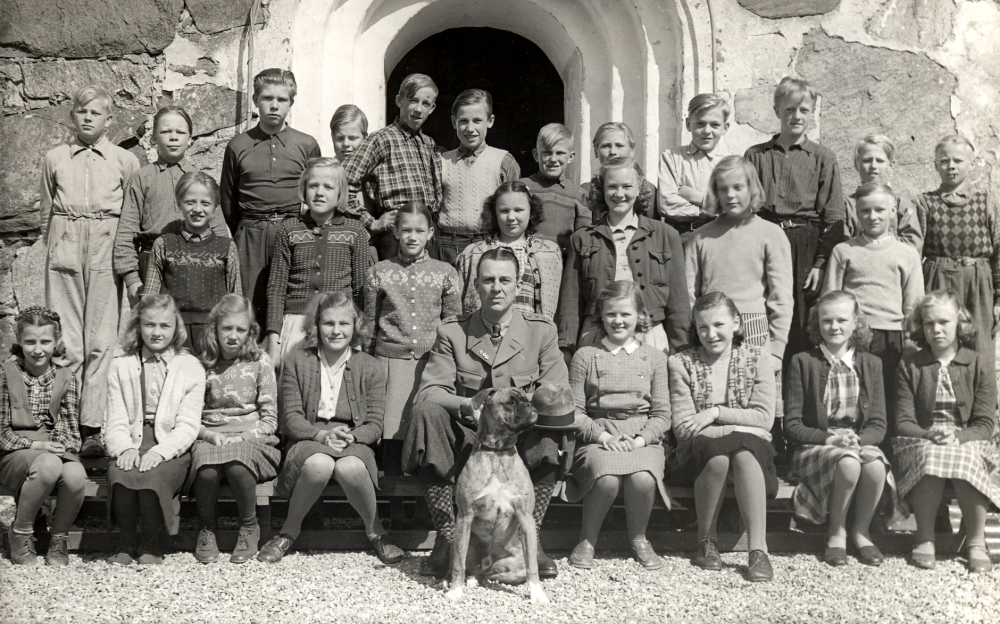Läraren Gösta Sahlin med skolbarnen i Össeby-Garns kyrkskola.  Klassen har samlats vid ingången till Össeby-Garns kyrka.  I bakre raden fr v Hans Gustavsson, Rolf Degling, Karl-Gustaf Spetz, Yngve Alstermark, Bengt Karlsson, Tord Wester, Olle Pettersson, Erik Forsberg, Ture Rödin, Karl-Erik Johansson, Holger Dahlbeck och Rune Jansson.  Mellanraden fr v Arne Johansson, Ingegerd Karlsson, Margareta Balck, Birgitta Lindell, Kerstin Balck, Britt Pettersson och Leif Johansson.  Sittande fr v Marianne Lundberg, Kerstin Johansson, Kerstin Stjärnholm, Gunilla Andersson, läraren Gösta Sahlin med en hund, Ella-Görel Eriksson (Wettervik), Ulla Franzén och Ella Gustavsson