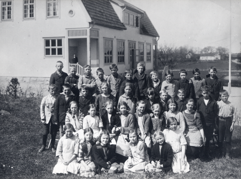 Skolbarnen samlade utanför Hjälmsta skola Vita skolan som uppfördes 1910. I bakgrunden syns den gamla arrendatorsbyggnaden vid Vallentuna prästgård samt alldeles t h om skolan skymtar Vallentuna kyrka. På trappan står en kvinna som kan vara lärarinnan Edit Lindström