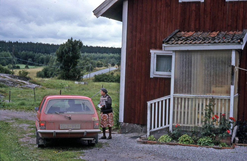 Lantbrevbäring i Vågsjö;  Elsa Wanzelius i Vågsjö, Täby, samtalar med lantbrevbärare Mikael Mattsson som sitter i bilen. Elsas man arbetade hos Häggberg som hade gården tvärs över vägen. Den blivande  Arningeleden ligger nu där de kortare ljusa träden möter de längre mörka träden.