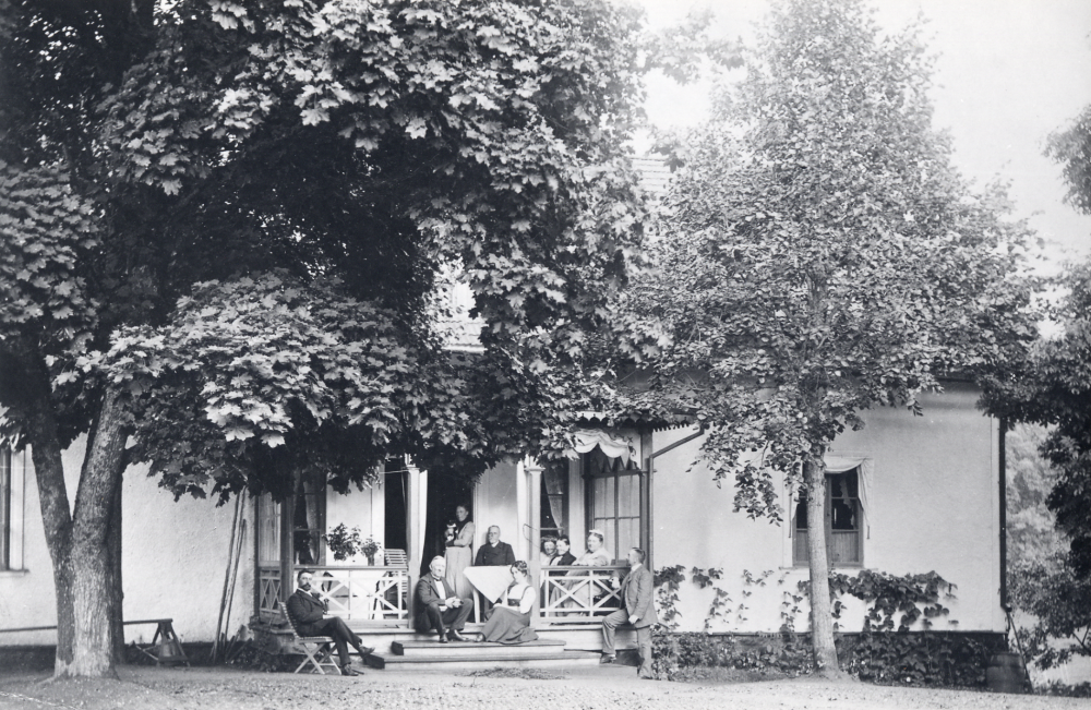 På verandan till Össeby-Garns prästgård sitter kyrkoherde Karl Hedén vid bordet och hans hustru Ella Hedén står i dörren. Övriga personer är familjen Nyman