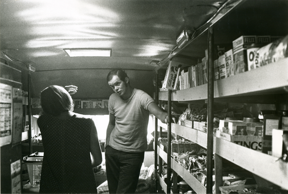 Arne Karlsson, som kör livsmedelsbussen från Frösunda handel, pratar med en kund. Interiör från bussen med varor på båda sidor.
•Fotografiet ingår i bildserien ”Vardagsliv i Vallentuna kommun” av Rachael Gough-Azmier & Gunilla Blomé, Haga Studios HB.