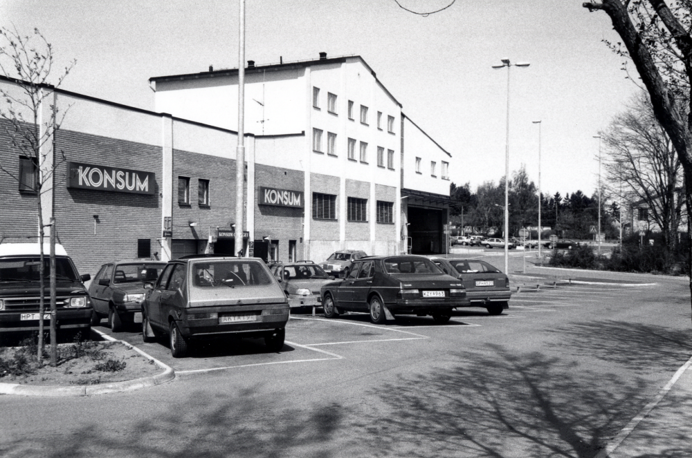 Konsumhusets baksida sett från Banvägens busshållplats med bilparkering i förgrunden och Konsumgaragets infart samt Konsumbutikens lastintag