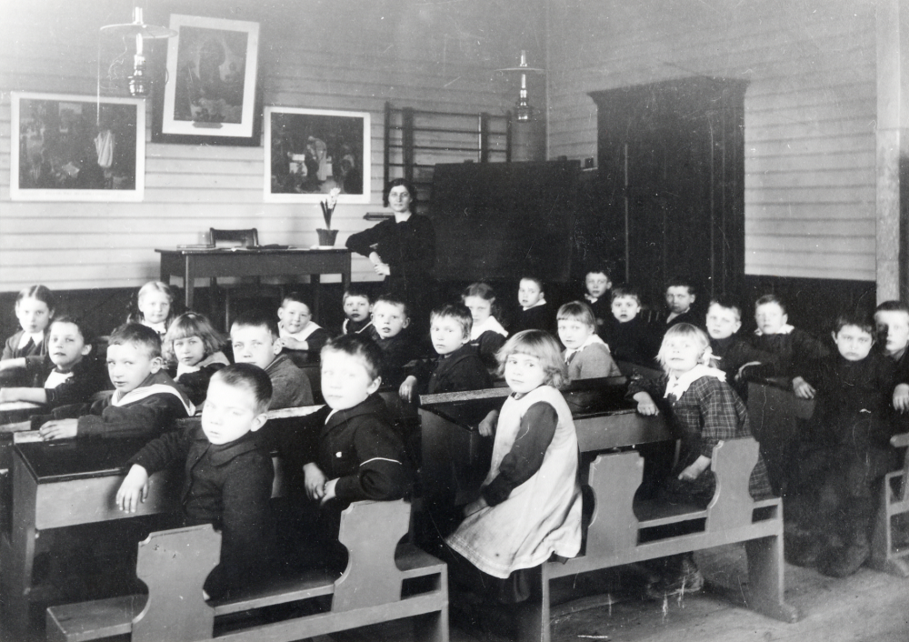 Interiör  från  Össeby-Garns kyrkskola  med lärarinnan  Nanny Skoglund  tillsammans med skolbarnen. I taket hänger två  fotogenlampor